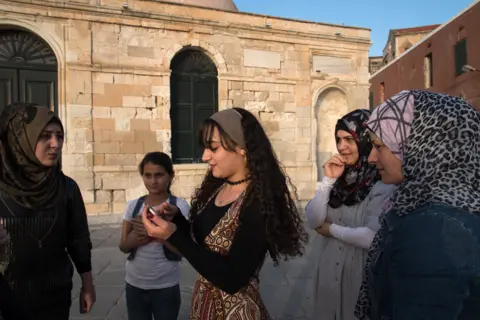 BBC Esraa Afandaki, 16, in the center, along with her sister Fatima,21, Reem Tarzalaki,11, Yasmin Afandaki,32,and Faten Tarzalakis, 36 go for an afternoon walk at Chania harbor. Behind them lie Giali Tzamisi, an imposing mosque in the harbor, that used to be one of the earlier buildings of the Ottomans in Crete. It is called Giali Tzamisi, which means "The Seaside Mosque", because it was built next to the sea.
