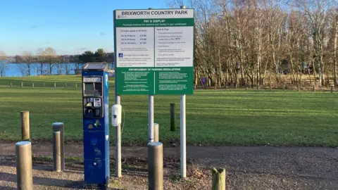 BBC Brixworth Country Park's current tariff displayed on a board next to a parking metre.