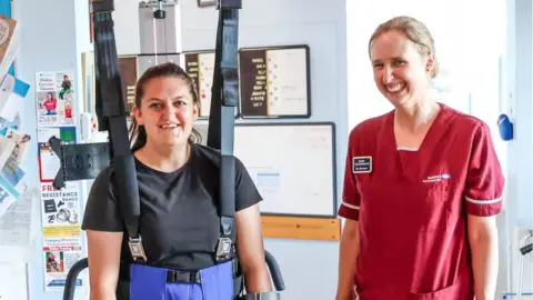 Spencer Mulholland Megan is on the left being held up standing using hoist straps and smiling. Elly is on the right in a red uniform smiling at the camera