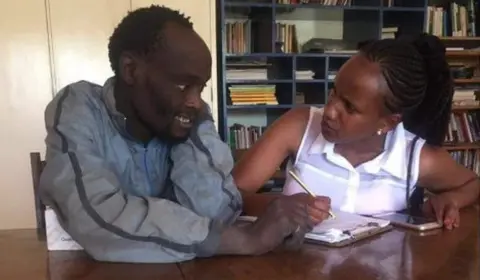Wanja Mwaura Wanja and Hinga sit on a desk indoors as she teaches him from a book. Hinga is smiling.