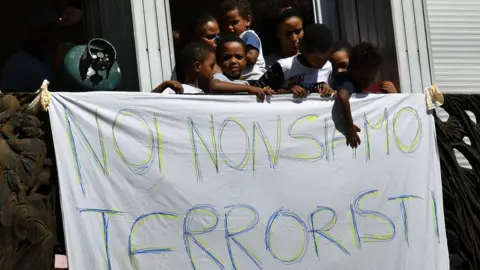 Getty Images Refugees and asylum seekers protest against their eviction near gas cylinders and a banner reading 'We are not terrorists' on 23 August 2017