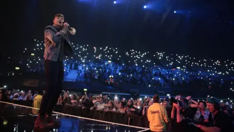 Kieron McCarron/BBC Man with short dark hair sings into a microphone on a stage. Audience holding lights.