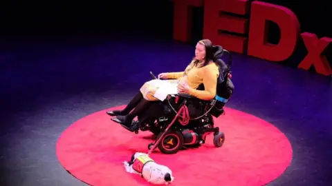 Lucy Watts Lucy Watts speaking at a TedX event with her support dog Molly at her side