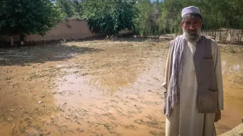 BBC Mohammad Rasool stands in front of a field with crops covered in muddy brown water