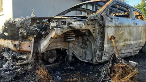 Matt Marvel/BBC The burnt out remains of a car destroyed in a suspected arson attack. The bonnet is almost entirely gone, parts of the engine are on show an a wheel is missing. A remnant of the wheel is on the ground next to the car shell.