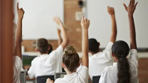 Getty Images A group of pupils in a classroom seen from behind. All of the pupils have white shirts on, and all of them have their hands raised as if to answer a question