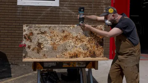 Chris Hoare A man is wearing a mask and working on a wall using electrical equipment