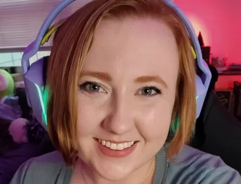 Actually Arcane Ashlyn is smiling, wearing light purple gaming headphones which emanate a green light from the ear cups. The headset covers her reddish hair, which is about chin-length. She's in a room which appears to have gaming memorabilia just visible in the background. One corner is bathed in a soft red light.