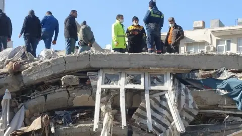 BBC People stand on top of rubble. An intact window is visible in the layers of rubble.