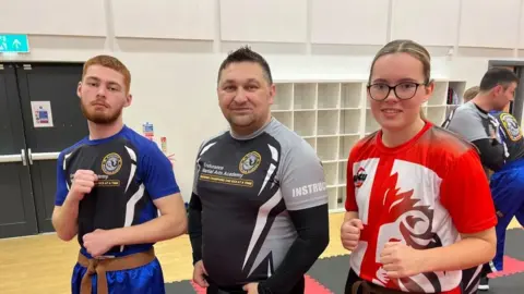 Ollie Conopo/BBC Young man with ginger beard in fighting pose, older man with slight beard and teenage girl in fighting pose with red top and glasses