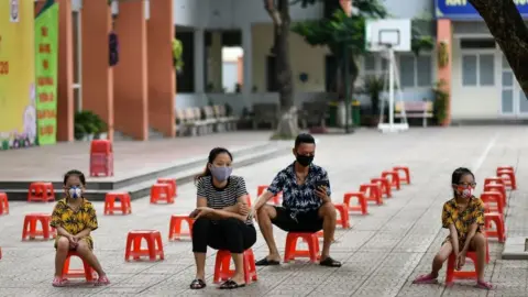 Getty Images A family sit on plastic stools as they wait to be tested at a makeshift rapid testing centre as Vietnam records a rise in cases of the COVID-19 coronavirus in Hanoi on July 31, 2020.