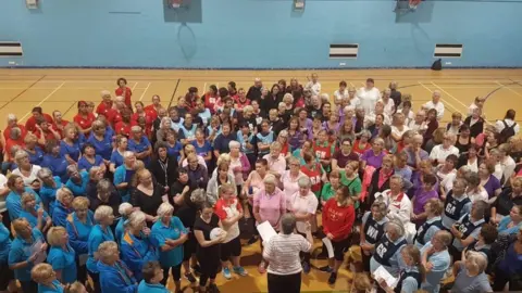 BBC Ms Squires stands at the front of a crowd in a sports hall, which are in a semi-circle shape around her