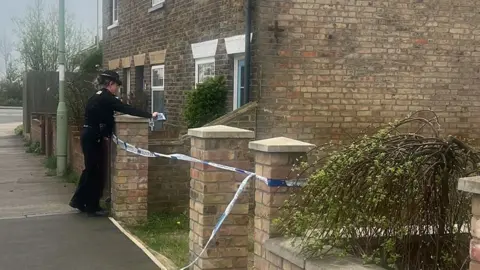 Lauren Fox Police officer tying police tape around a brick gate pillar