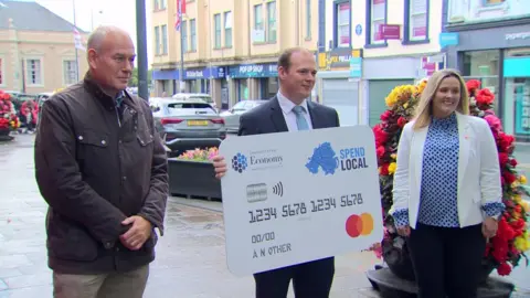 Gordon Lyons holds a giant version of the high street voucher on a street in Carrickfergus with two people next to him