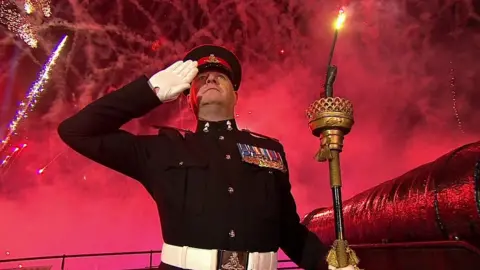 Soldier saluting while holding torch next to cannon with fireworks in background