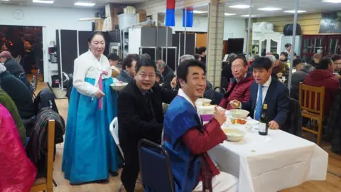 Je Seung Lee/BBC Woman in traditional Korean outfit stands at the top of a table. Men are seated at the table with food in front of them. They are all smiling at the camera.