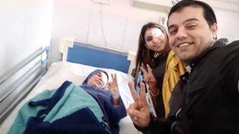 Mohammad Farzi Elahe Tavokolian lying in a hospital bed with Mohammad Farzi (R) and Kosar Eftekhari (C) next to her