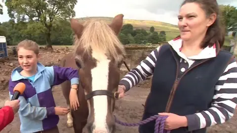 A woman and her daughter standing either side of a horse with a microphone being pointed at the girl