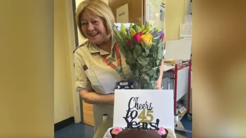Bristol Royal Hospital for Children Image of Sue Sheriton holding a bunch of flowers. A card reading Cheers to 45 Years! is in front of her, along with a chocolate cake.