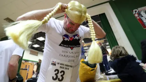PA Malcolm Pernet puts on a wig as he prepares to carry Kate Barfield in the annual UK Wife Carrying Race