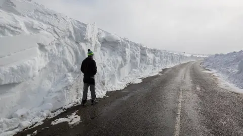 Getty Images A man stands beneath a huge snow drift on a road in the Pennines on March 20, 2018 in Allenheads, England