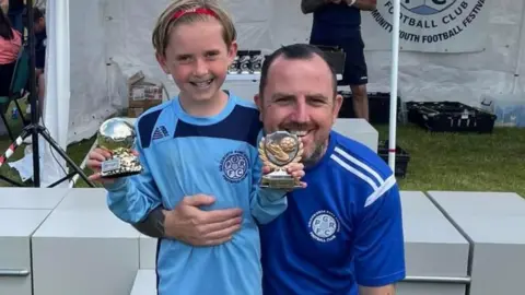 Family photo Matthew Evans with his arm around son Ollie holding a football trophy