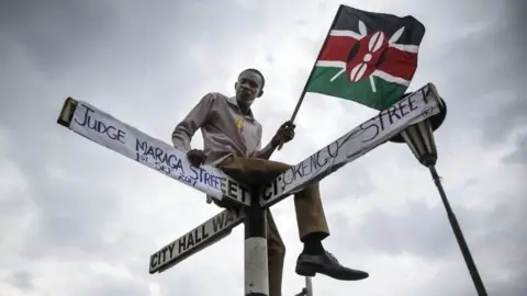 EPA A supporter of The National Super Alliance (Nasa) opposition coalition and its presidential candidate Raila Odinga sits on top of a street sign post that has been relabeled "Judge Maraga Street", referring to Chief Justice David Maraga,