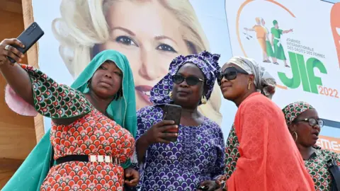AFP Women taking selfies in front of a International Women's Day poster showing the Ivorian first lady, Abidjan, Ivory Coast - Tuesday 8 March 2022