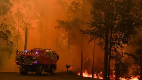 EPA A fire truck near a raging fire in Werombi, south-west of Sydney, against a bright orange sky