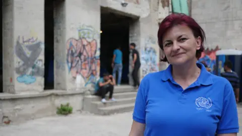 BBC Amira, in a blue and white collared t-shirt, stands in front of graffiti-marked pillars between which men loiter