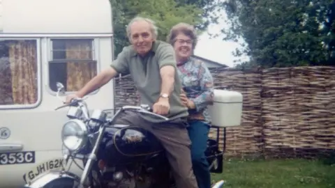 Peter Daines Mano on motorbike with woman riding pillion beside a caravan