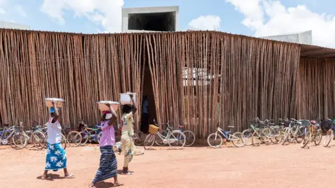 Iwan Baan Women walking outside Lycée Schorge Secondary School. There are bikes in the background. The building is made up of tall brown sticks.