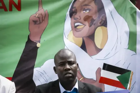 EPA A man looks ahead at the camera. Behind him is a depiction of a young woman wearing large gold earrings and white fabric draped over her head and shoulders, her armed outstretched.