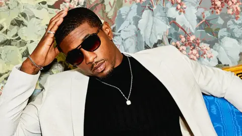 ASOS Love Island star Ovie Soko models his ASOS collection wearing dark sunglasses, a white suit and a dark polo neck sweater