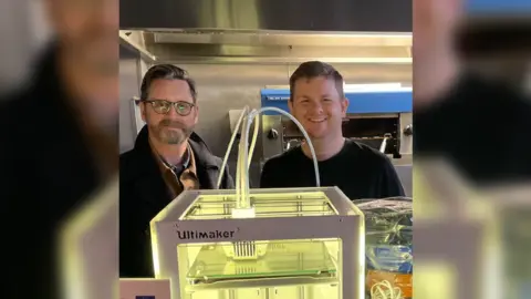 Gary Buller Two men stand in a workshop. In front of them is a large machine.