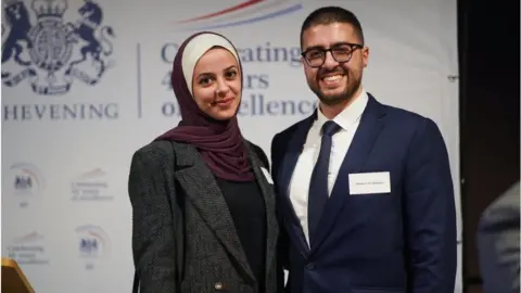 FCDO Dr Maisara Al Rayyes and his fiancée pose for a photo in front of a Chevening Scholarships branded backdrop