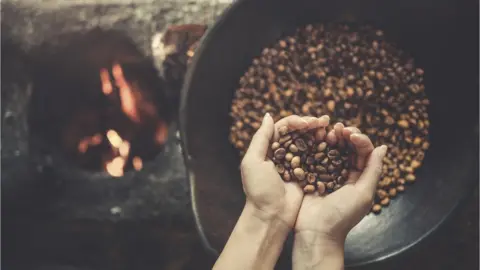 Getty Images Female hands holding freshly roasted coffee beans above a bowl next to a fire.