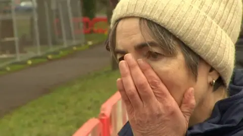 BBC A female protestor shares an expression of disbelief as she looks on with her right hand on her face as the trees are cut down