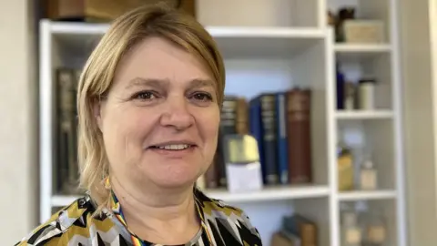 Lesley Self looks into the camera in front of a bookshelf of old books and vintage medicine bottles