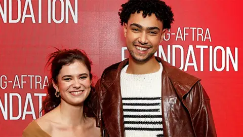 Getty Images Alison and Archie, standing next to each other with a red background. Alison is smiling, wearing a brown outfit, while Archie, who is taller, is wearing a white and black patterned jumper under a brown leather jacket.