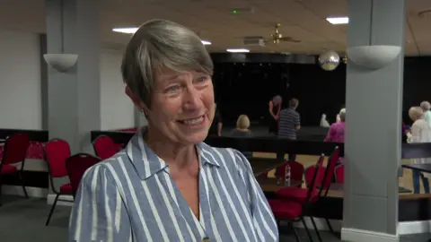 A lady in a striped blouse stands in a hall where others are doing Tai Chi