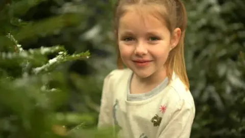 Ellie Hawkes Image of Lyra Cole standing amidst Christmas trees looking into the camera