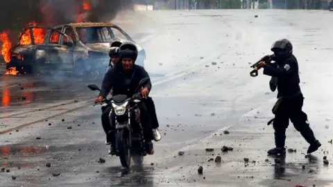 Reuters A riot police officer fires his shotgun towards two men during on a motorcycle on May 28, as the remains of a car burn brightly in the background