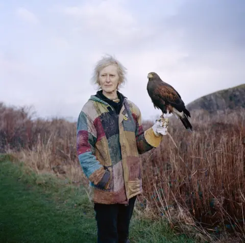 Jon Tonks A woman in a colourful jacket stands in the countryside and holds a hawk