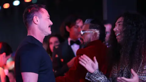 Getty Images Cole Walliser speaking to actor Luke Evans on a red carpet in Cannes, France, in 2023. Cole (right) is a Canadian man in his 40s with long curly brown hair past his shoulders. He has brown eyes and a short moustache. He has his arms open towards Luke, a white man in his 40s with cropped brown hair and a thick moustache. Cole wears a black and white patterned shirt while Luke is styled in a black T-shirt. Behind them are other red carpet guests and reporters.