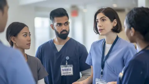 Getty imagines a group of five healthcare workers having a conversation in a hospital. Three women and two men wear blue scrubs and ID badges.