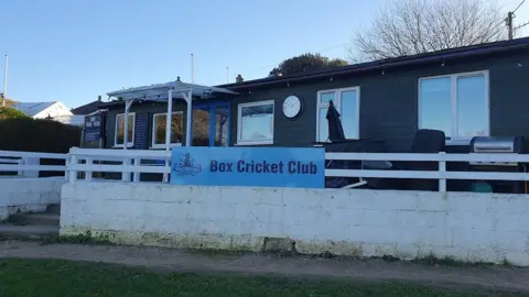 BBC A one storey wooden building with a sign for Box Cricket Club and a blue sky