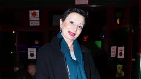 Shutterstock A white woman, with short black hair, tilting her head wearing a blue scarf and black fleece. The background is dark with stickers on the brown wall.