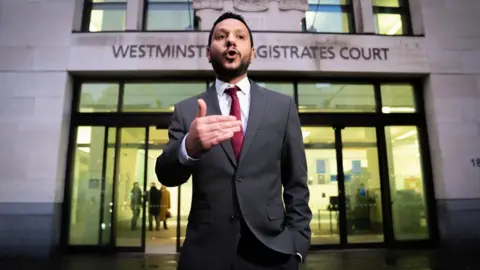 PA Media Sayed Ahmed Alwadaei, wearing a dark suit and red tie, speaking to the media outside Westminster Magistrates' Court in central London