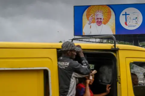 Getty Images A billboard shows an image of Pope Francis in Kinshasa as passengers travel on a minibus past it.